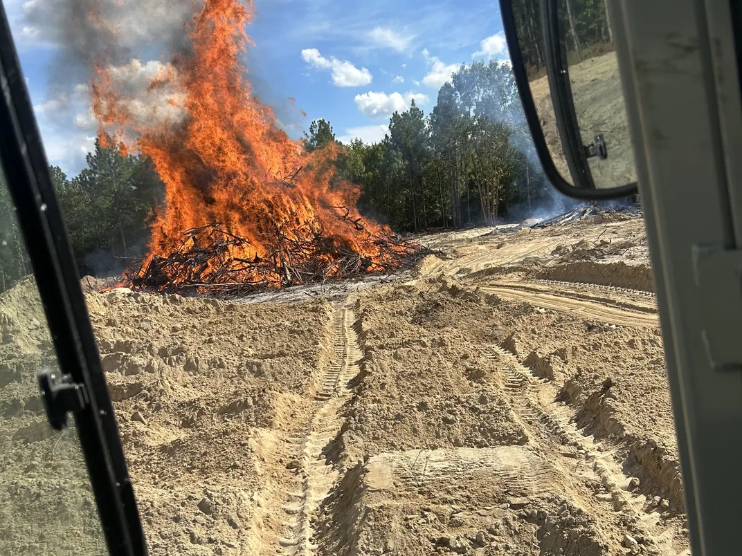 Pile of cleared brush burning on a sandy lot seen from the excavator cab, lot clearing