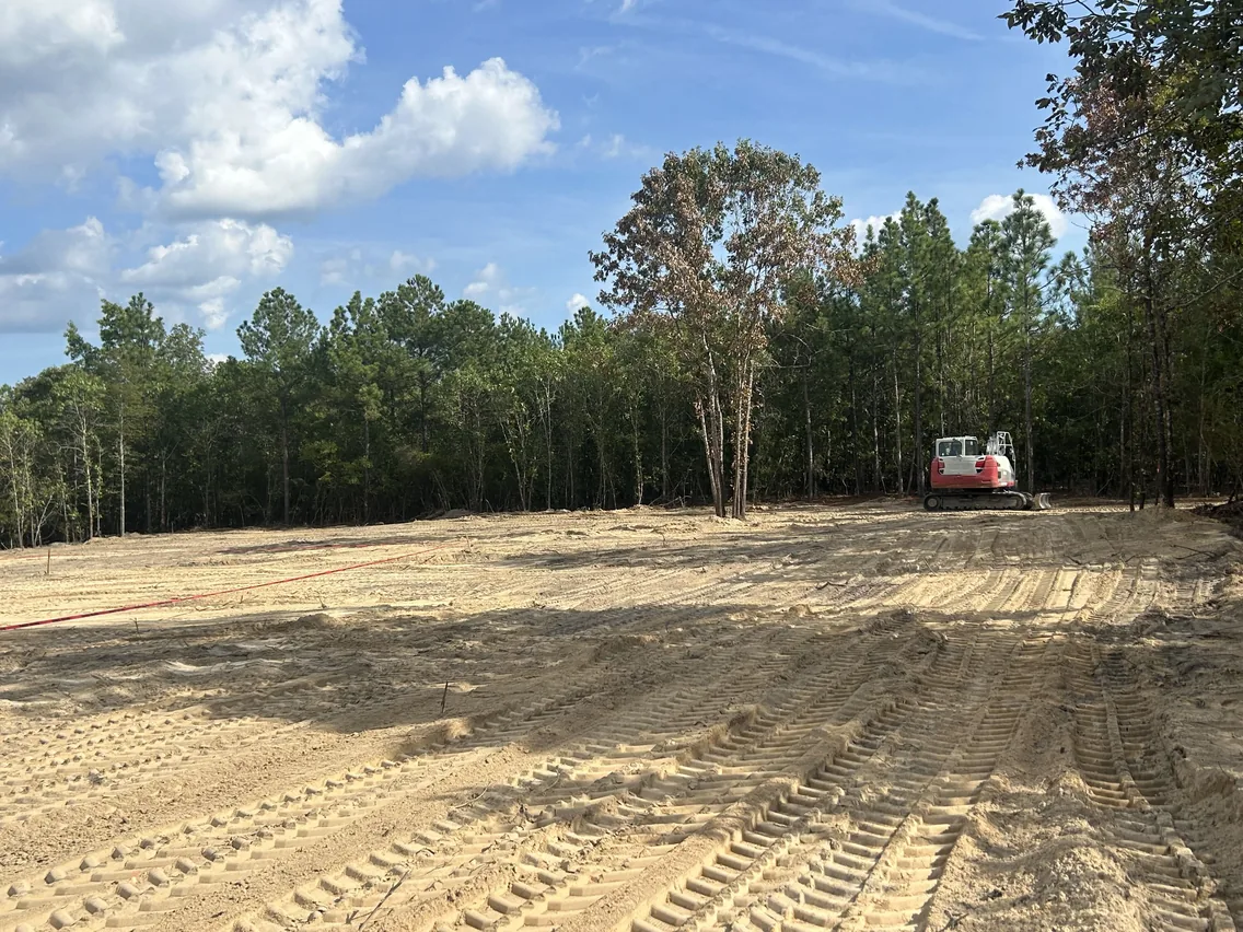 Cleared homesite lot with fresh track marks and an excavator by the pine tree line, lot clearing