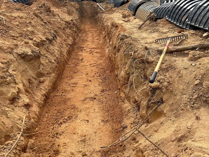 Open trench dug in red clay with chambers and a rake on the bank, septic system repair