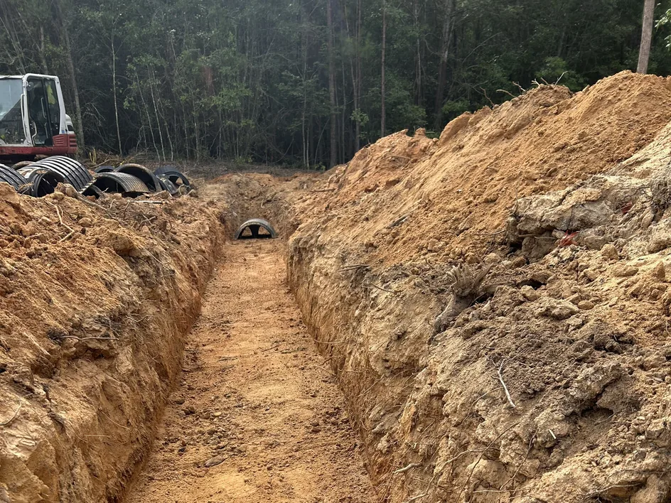 Dug trench with a chamber set at the far end and an excavator by the woods, septic system repair