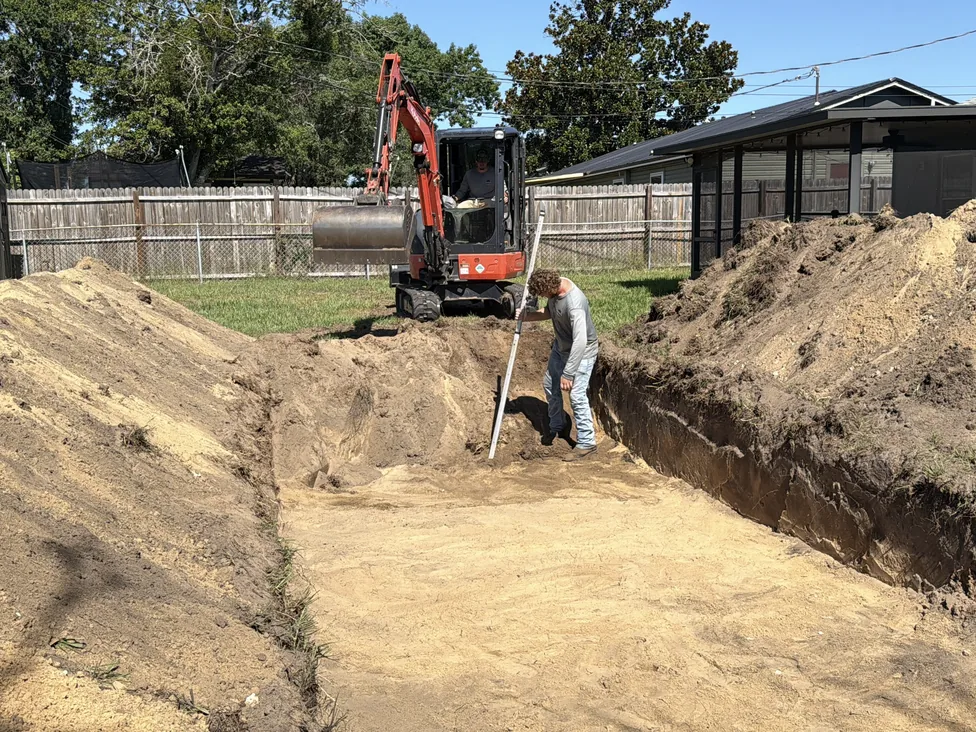 Mini excavator and a worker with a grade rod digging a new drain field bed in a fenced backyard, septic system replacement