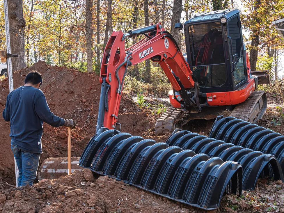 Mini excavator and a worker beside a row of drain field chambers by a fresh trench, septic system replacement